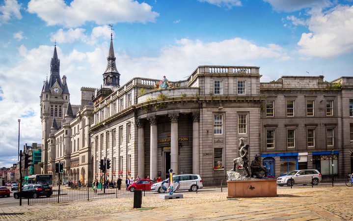 A grand historic building with columns and domed architecture, situated on a cobbled street with parked cars, pedestrians, and sculptures in front, under a partly cloudy sky.