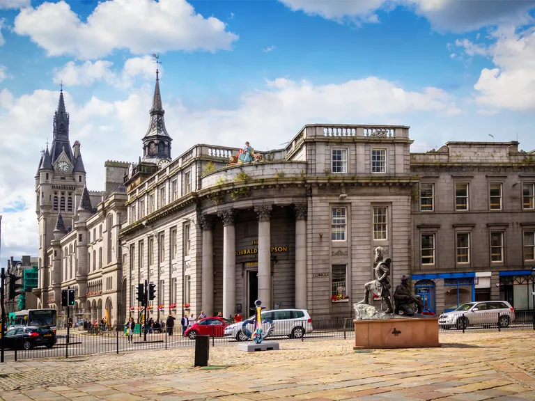 A grand historic building with columns and domed architecture, situated on a cobbled street with parked cars, pedestrians, and sculptures in front, under a partly cloudy sky.