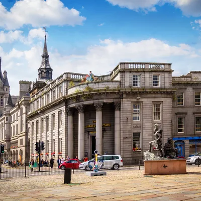 A historic building with classical architecture, columns and statues, set against a bright blue sky with scattered clouds, busy street with cars and pedestrians.