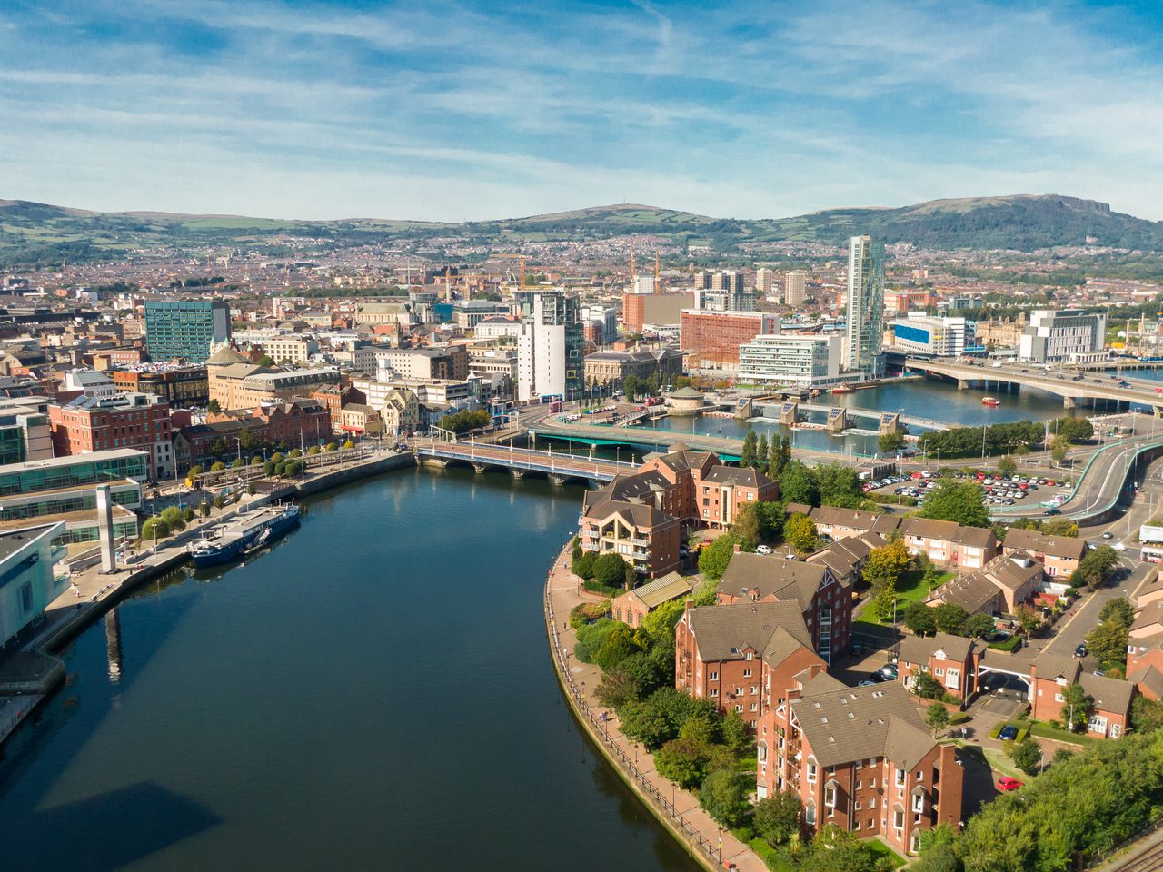 Veduta aerea di un fiume che attraversa una città moderna con edifici alti, case residenziali e un ponte, circondata da colline verdi sullo sfondo.
