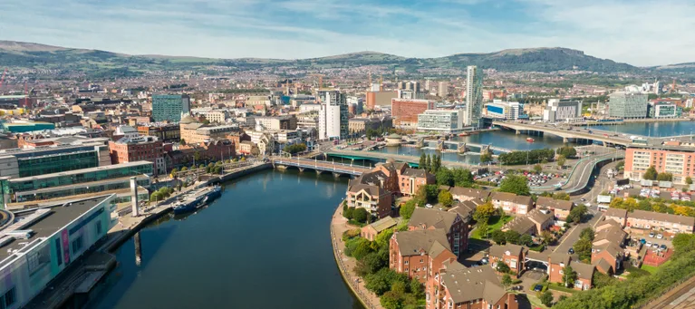 Aerial view of a city with a river running through, surrounded by modern buildings, residential houses, and hills in the background on a clear day.