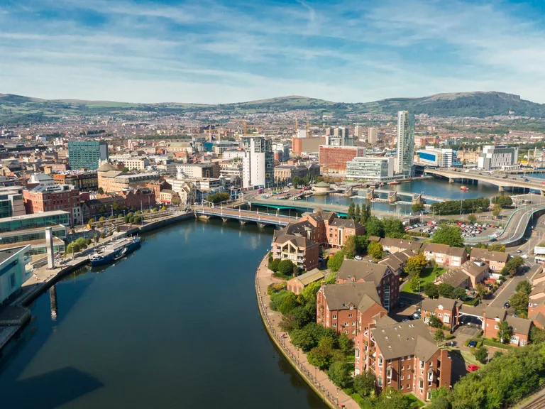 Aerial view of a city with a river running through, surrounded by modern buildings, residential houses, and hills in the background on a clear day.