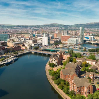 Aerial view of a cityscape with a river, modern buildings, bridges, and a mix of residential and commercial areas, set against rolling hills in the background.
