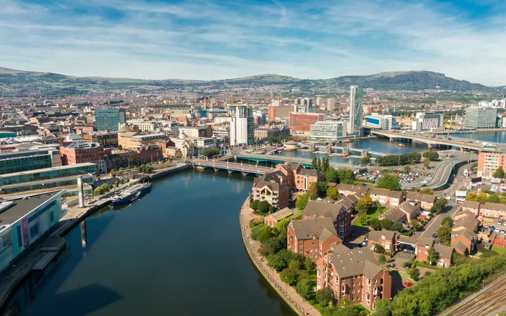Aerial view of a city with a river, modern buildings, residential houses, bridges, and mountains in the background under a blue sky.