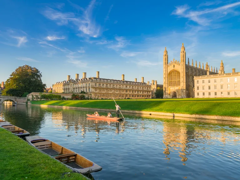 People rowing a boat on a river with historic buildings, including a cathedral, in the background under a clear blue sky.