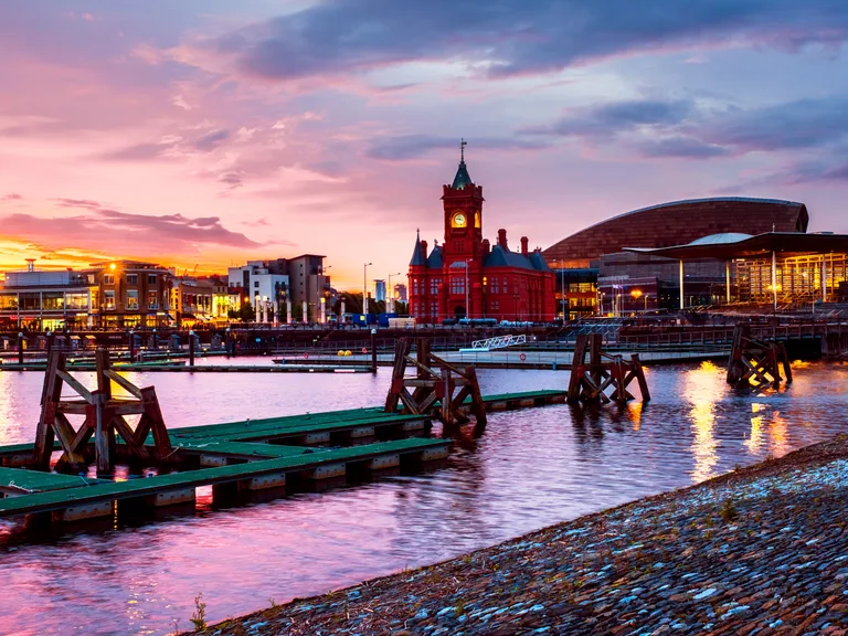 Colourful cityscape at sunset with a red clock tower over modern buildings, a waterway with wooden docks, and a cloudy sky.