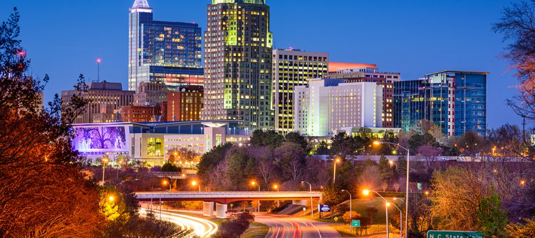 Night view of downtown high-rise buildings illuminated, with light trails on a curved highway in the foreground and trees along the road.