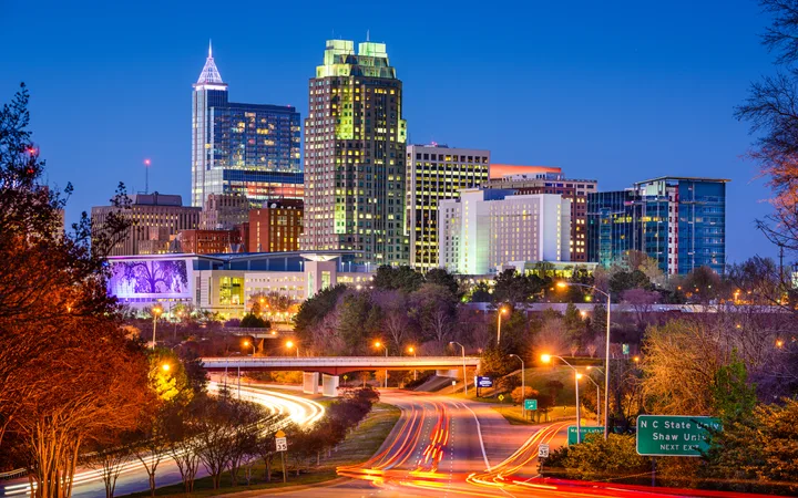 Nighttime city skyline with illuminated skyscrapers, busy roads with light trails, and trees in the foreground.