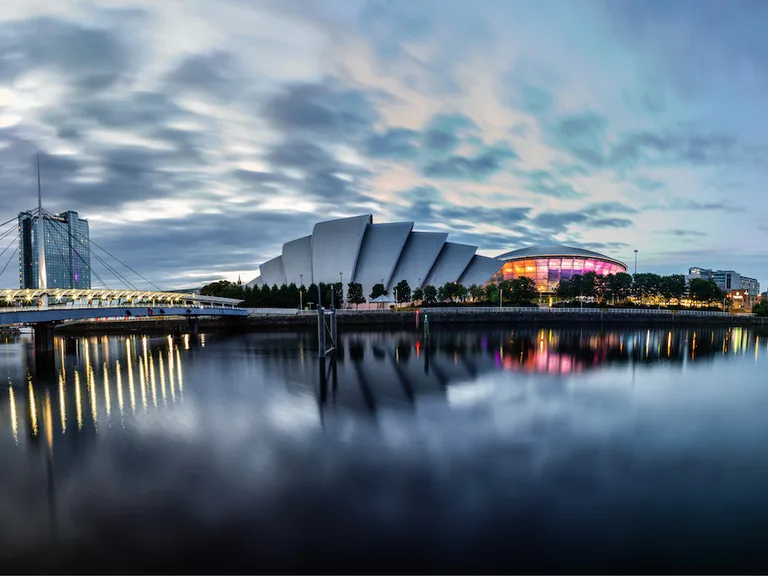 City skyline at dusk with modern buildings, a bridge over calm water, and colorful reflections on the surface. Sky filled with clouds.