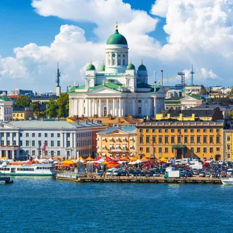 View of Helsinki waterfront with the Uspenski Cathedral on a bright, partly cloudy day, bustling market stalls, boats on the water, and city buildings in the background.