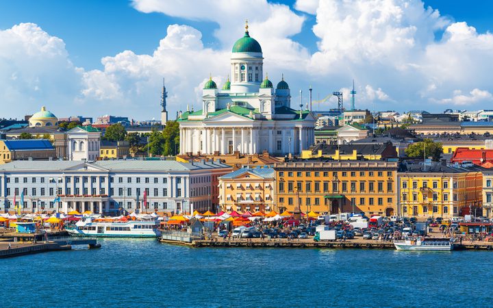 View of Helsinki waterfront with the Uspenski Cathedral on a bright, partly cloudy day, bustling market stalls, boats on the water, and city buildings in the background.