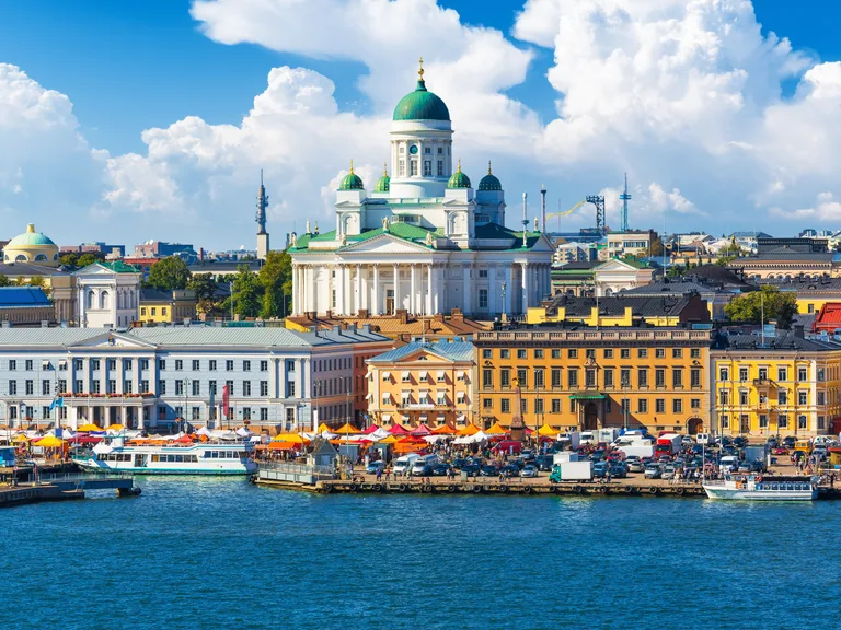 View of Helsinki waterfront with the Uspenski Cathedral on a bright, partly cloudy day, bustling market stalls, boats on the water, and city buildings in the background.