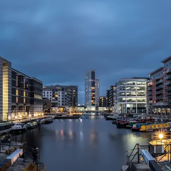 Veduta di un quartiere residenziale con edifici moderni, un porto e barche, al crepuscolo con luci accese e nuvole nel cielo.