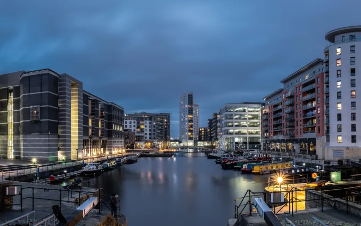 Bâtiments modernes au bord d'un canal urbain sous un ciel nuageux en soirée, avec des lumières reflétées dans l'eau et des bateaux amarrés.