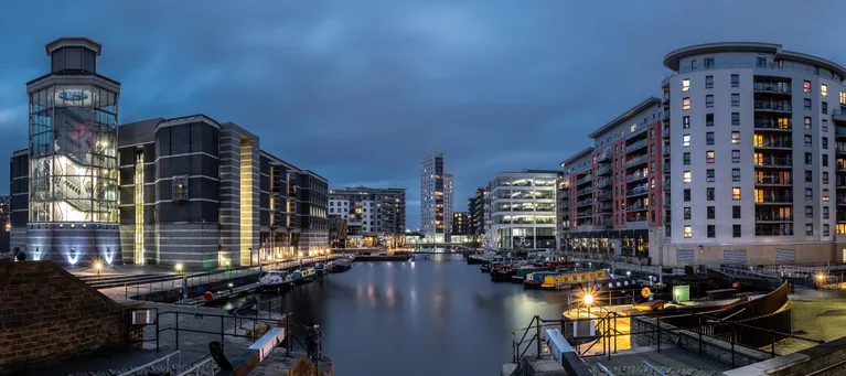 Modern cityscape with illuminated buildings and boats docked in a canal during dusk, under a cloudy sky.
