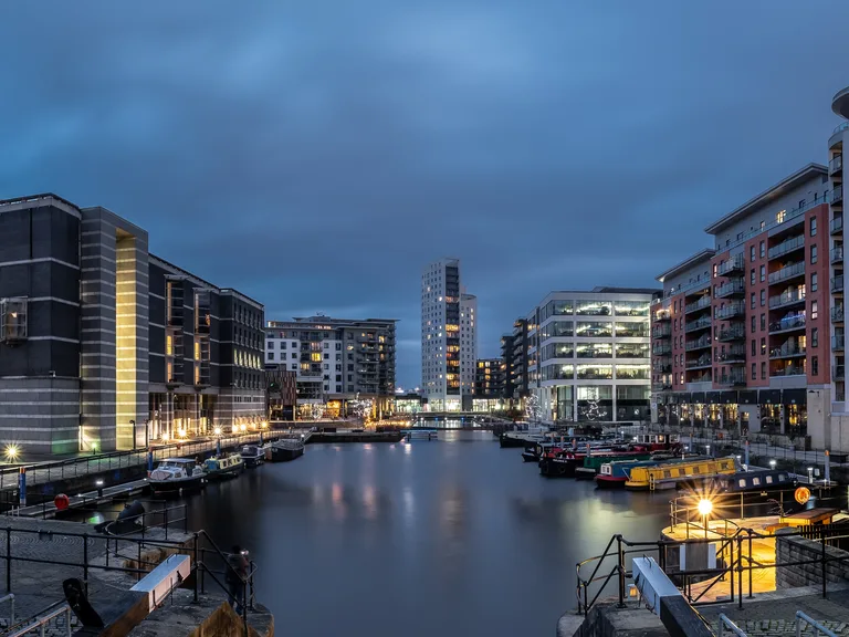 Nighttime view of a modern city harbor with illuminated buildings, boats docked along the waterfront, and a cloudy sky overhead.