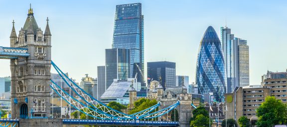 London skyline featuring Tower Bridge in the foreground and modern skyscrapers, including the Gherkin and the Cheesegrater, against a clear blue sky.