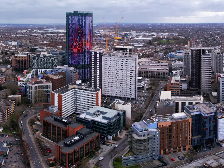 Aerial view of a city with modern skyscrapers, construction cranes and a mix of residential and commercial buildings under a cloudy sky.