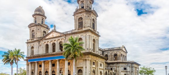 An old stone church with twin bell towers topped with crosses, surrounded by palm trees and a lush green lawn under a partly cloudy sky.