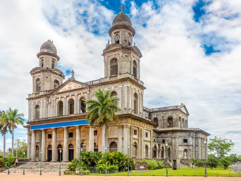 Chiesa storica con due torri, affacciata su un giardino con palme e lampioni, sotto un cielo nuvoloso.