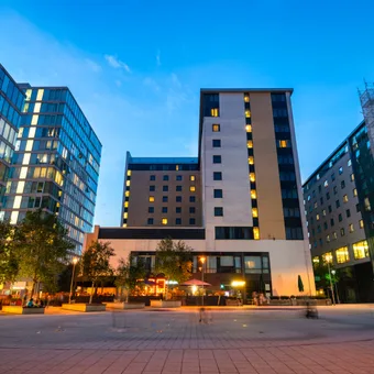 Modern cityscape with tall buildings, illuminated windows, a paved plaza, and trees against a clear evening sky.