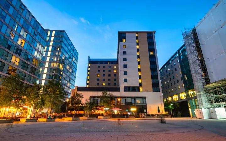 Modern cityscape with tall buildings, illuminated windows, a paved plaza, and trees against a clear evening sky.