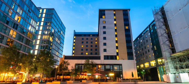 Modern cityscape with tall buildings, illuminated windows, a paved plaza, and trees against a clear evening sky.