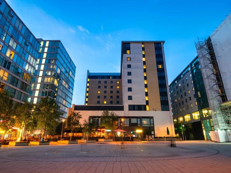 Modern cityscape with tall buildings, illuminated windows, a paved plaza, and trees against a clear evening sky.
