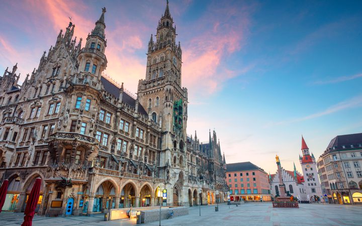 A historic Gothic-style building with ornate architecture and towers, set against a colorful evening sky in a spacious city square with few people.