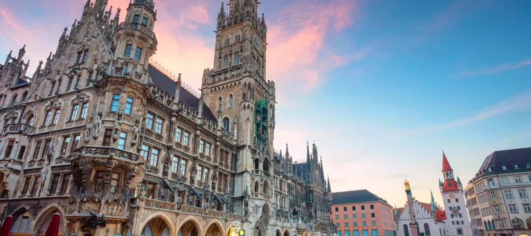 A historic Gothic-style building with ornate architecture and towers, set against a colorful evening sky in a spacious city square with few people.