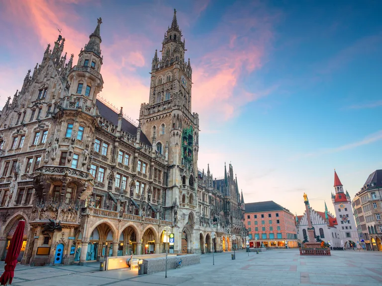 Historic gothic-style building with ornate towers and a central clock tower, set against a pink and blue sky at dusk, in a spacious city square.