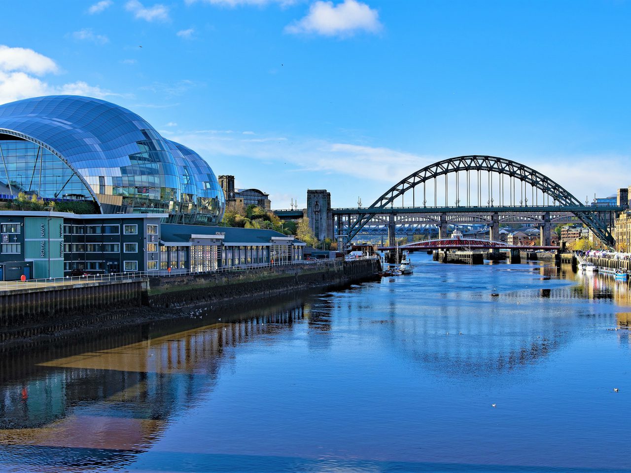Vue du pont et du bâtiment moderne en verre sur la rivière, sous un ciel bleu avec quelques nuages.