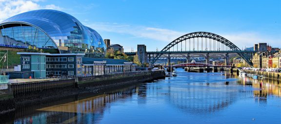 Modern glass building and arched bridge over a river under a bright blue sky with some clouds, surrounded by cityscape.