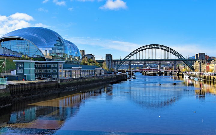 Modern glass-domed building beside a river with a bridge and historic buildings in the background under a bright blue sky.