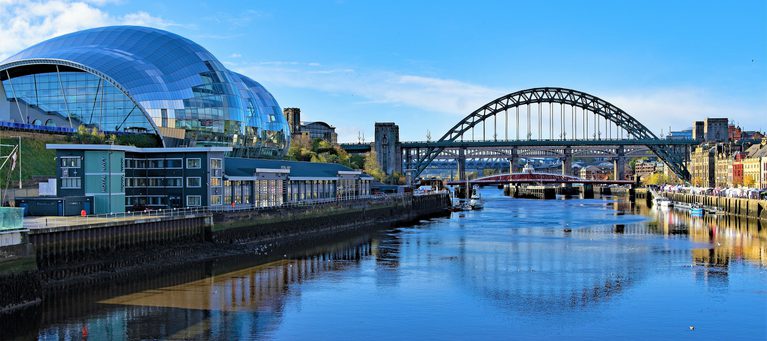 Modern glass-domed building beside a river with a bridge and historic buildings in the background under a bright blue sky.