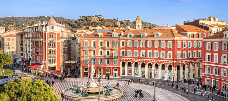 A vibrant city square with red buildings, a fountain with statues in the centre, and people walking on the streets under a clear blue sky.