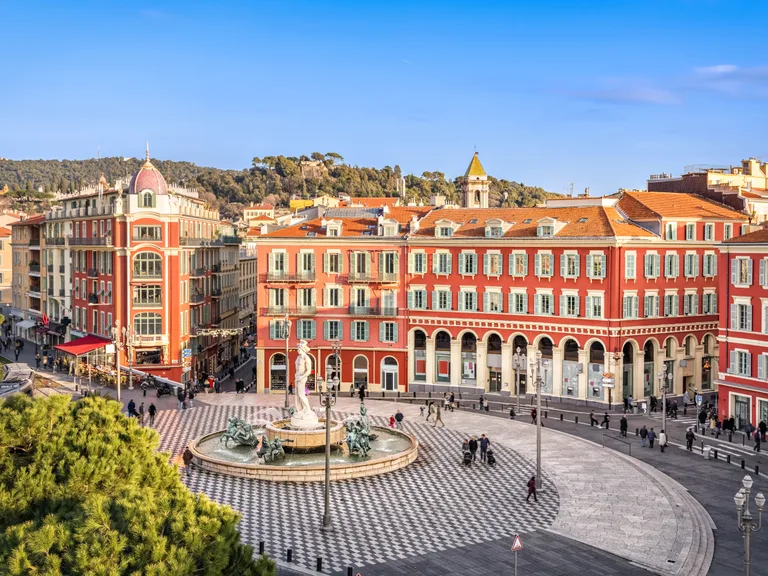 A vibrant city square with red buildings, a fountain with statues in the centre, and people walking on the streets under a clear blue sky.