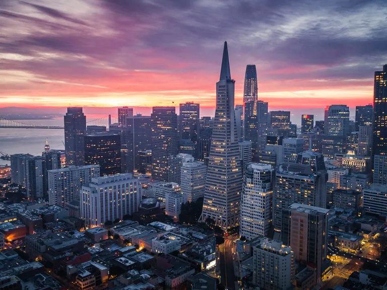 Sunset over San Francisco skyline with iconic buildings, Bay Bridge in the background, and colorful sky overhead, city lights beginning to shine.