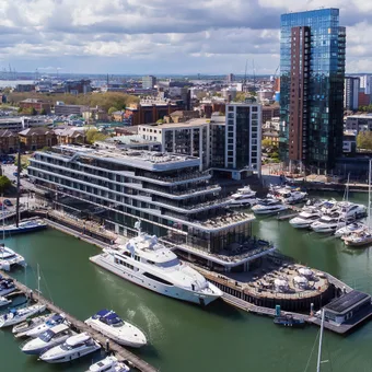 Aerial view of a harbour with yachts docked and modern high-rise buildings in a cityscape under a partly cloudy sky.