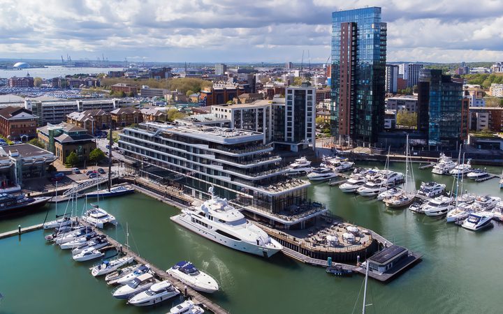 Aerial view of a harbour with yachts docked and modern high-rise buildings in a cityscape under a partly cloudy sky.