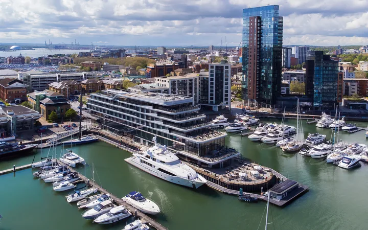 Aerial view of a harbour with yachts docked and modern high-rise buildings in a cityscape under a partly cloudy sky.