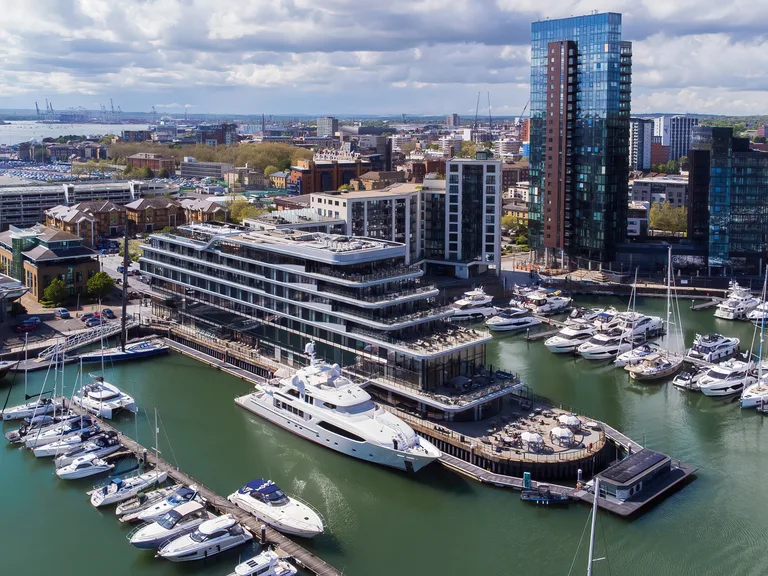 Aerial view of a harbour with yachts docked and modern high-rise buildings in a cityscape under a partly cloudy sky.