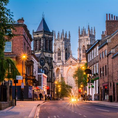 A street scene at dusk with historic brick buildings, a prominent cathedral in the background, and street lights illuminating the road.