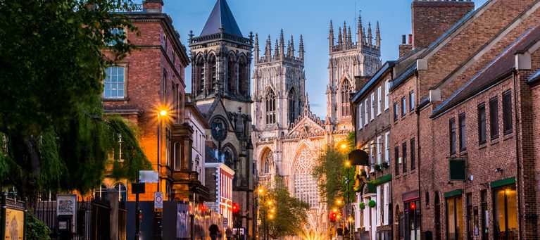 A quiet city street at dusk with historic brick buildings and a large Gothic cathedral in the background, illuminated by streetlights.