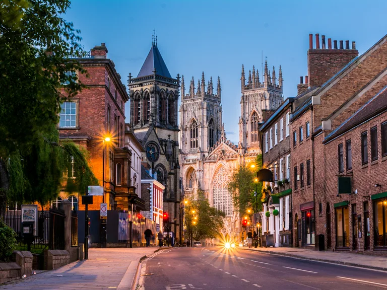 A street scene at dusk with historic brick buildings, a prominent cathedral in the background, and street lights illuminating the road.