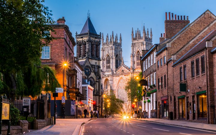 View of a city street with historic brick buildings, streetlights, and a large Gothic cathedral in the background during dusk.