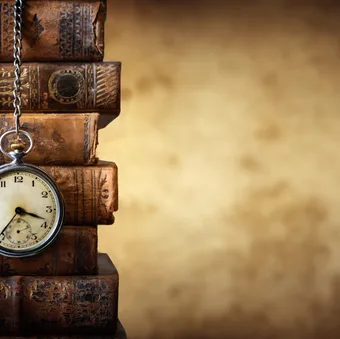 An antique pocket watch hanging from a chain on a stack of old, leather-bound books with ornate covers, against a blurred warm-toned background.