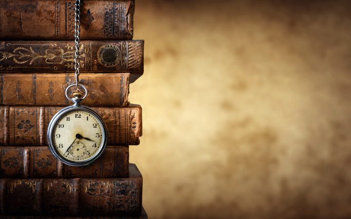 An antique pocket watch hanging from a chain on a stack of old, leather-bound books with ornate covers, against a blurred warm-toned background.