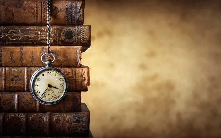 A vintage pocket watch hanging from a chain on a stack of old, worn leather-bound books against a blurred beige background.