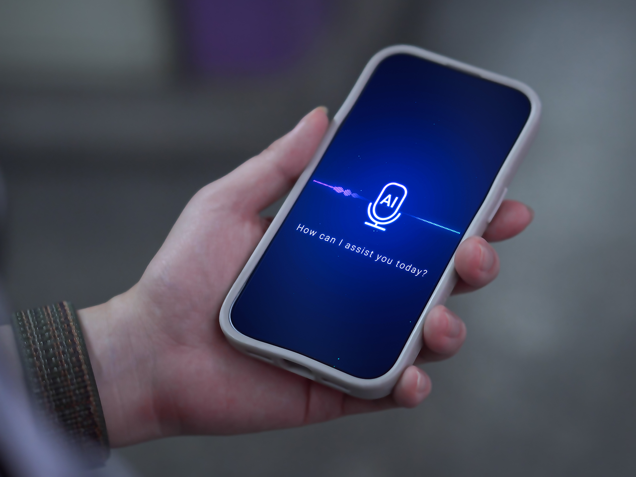 A hand holding a smartphone with a blue screen showing an AI microphone icon and the text 'How can I assist you today?'